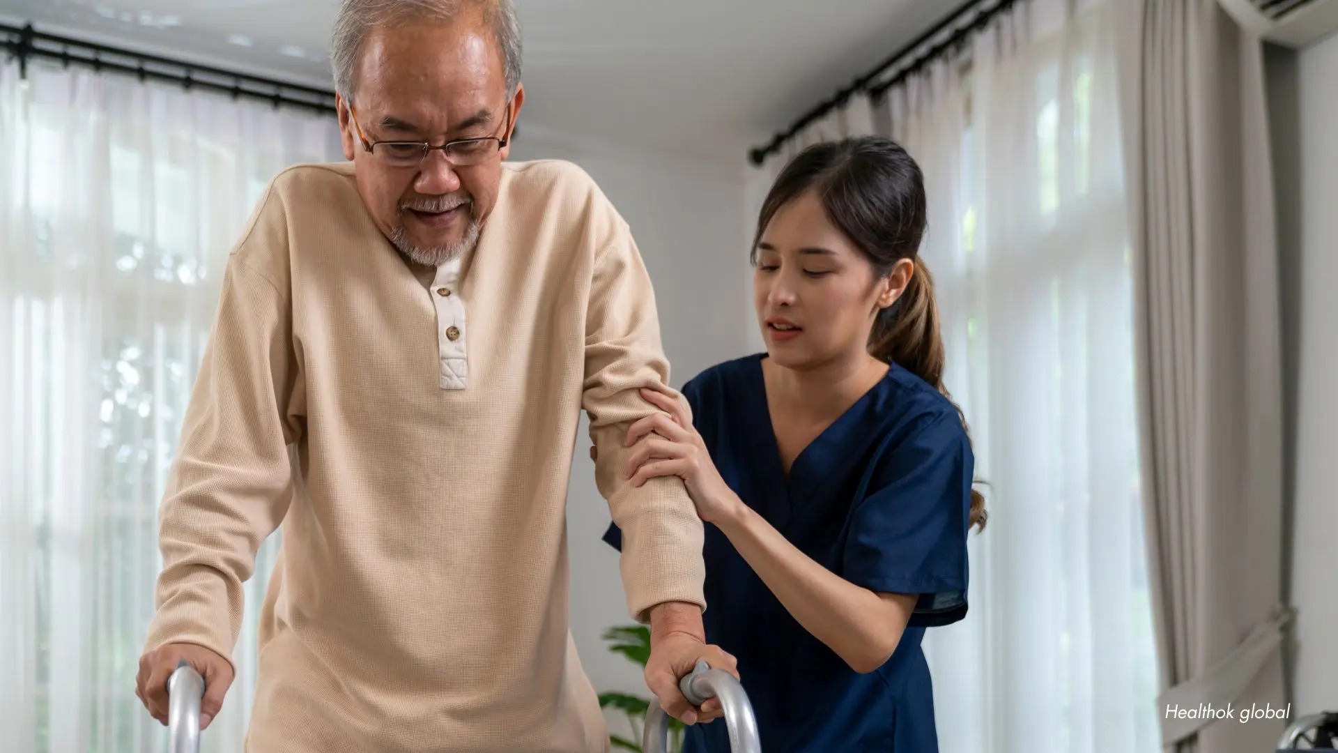 Elderly man using a walker assisted by a caregiver in a bright living room - support and elderly care concept.