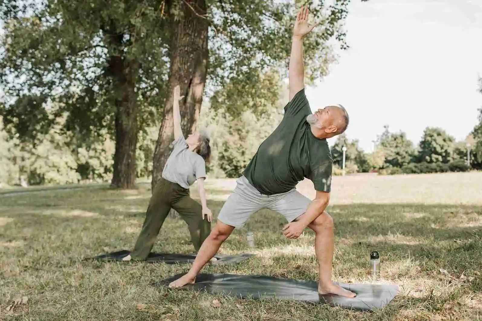 Elderly couple practicing yoga outdoors in a park, promoting physical fitness and mental well-being for seniors through gentle stretching exercises.