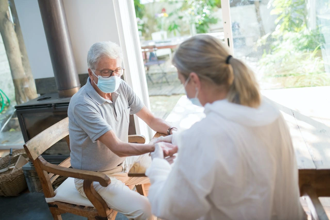 An elderly man wearing a mask receives care from a healthcare professional during a home consultation. The image emphasizes senior health, home-based care services, and COVID-19 safety measures.