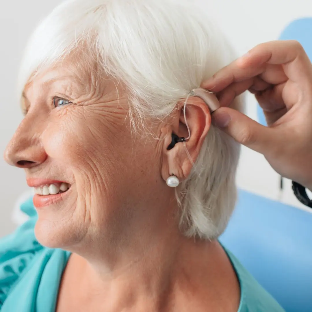 Elderly woman smiling while receiving assistance with digital hearing aid fitting at a healthcare clinic.