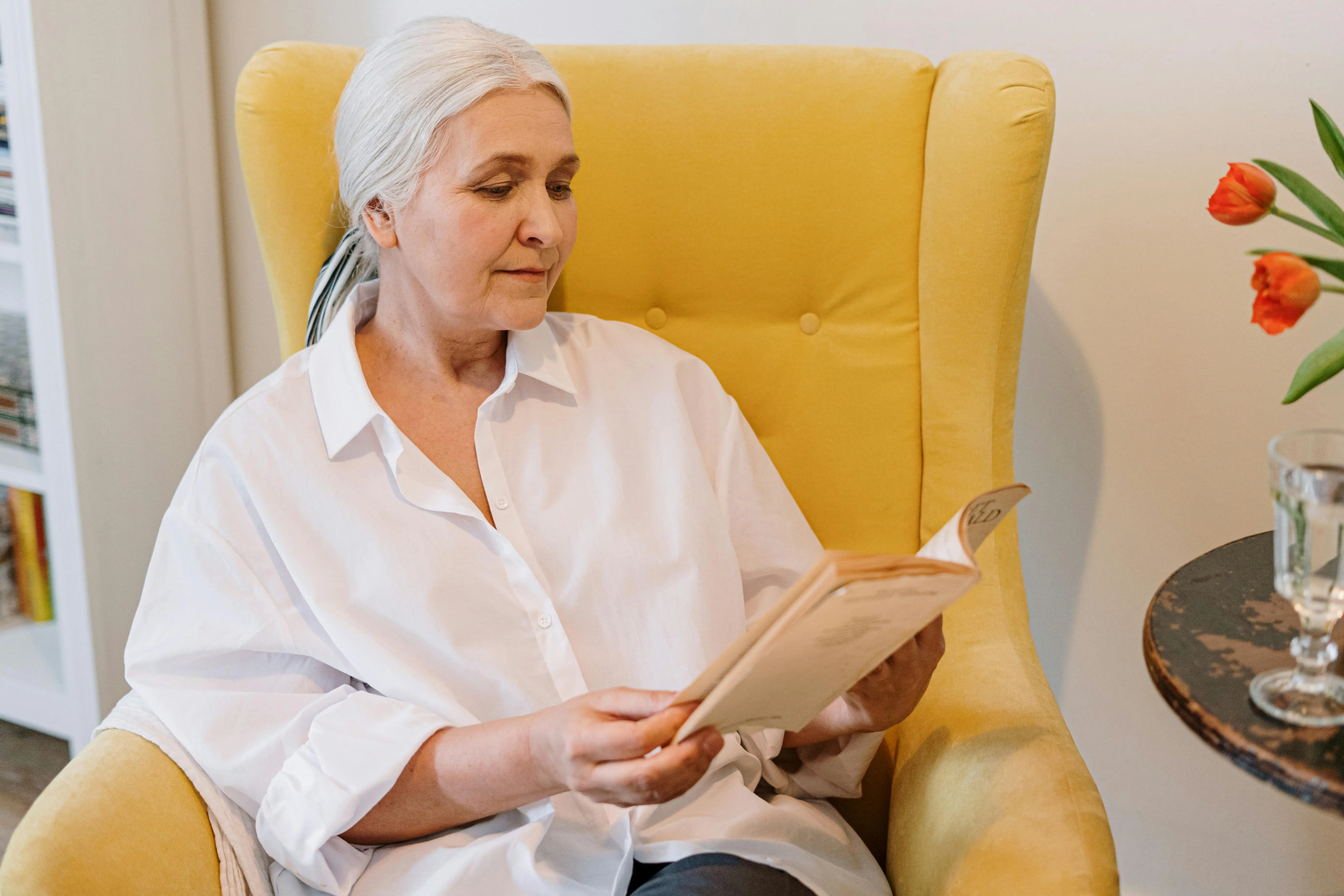 Elderly woman reading a book while sitting in a cozy yellow armchair, promoting senior mental health and relaxation activities.