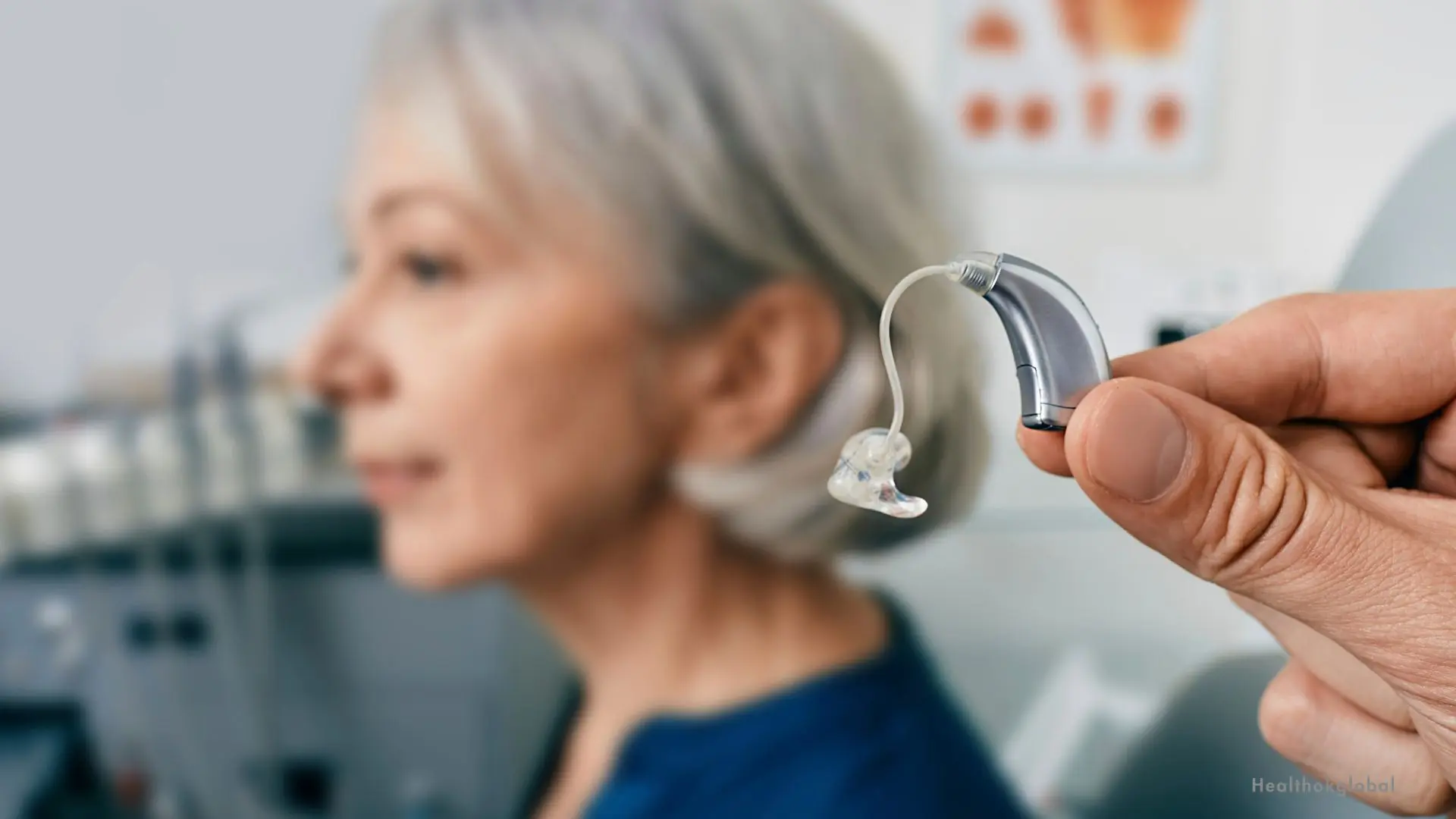 Close-up of a modern hearing aid held by an audiologist with a senior woman in the background during a hearing consultation.