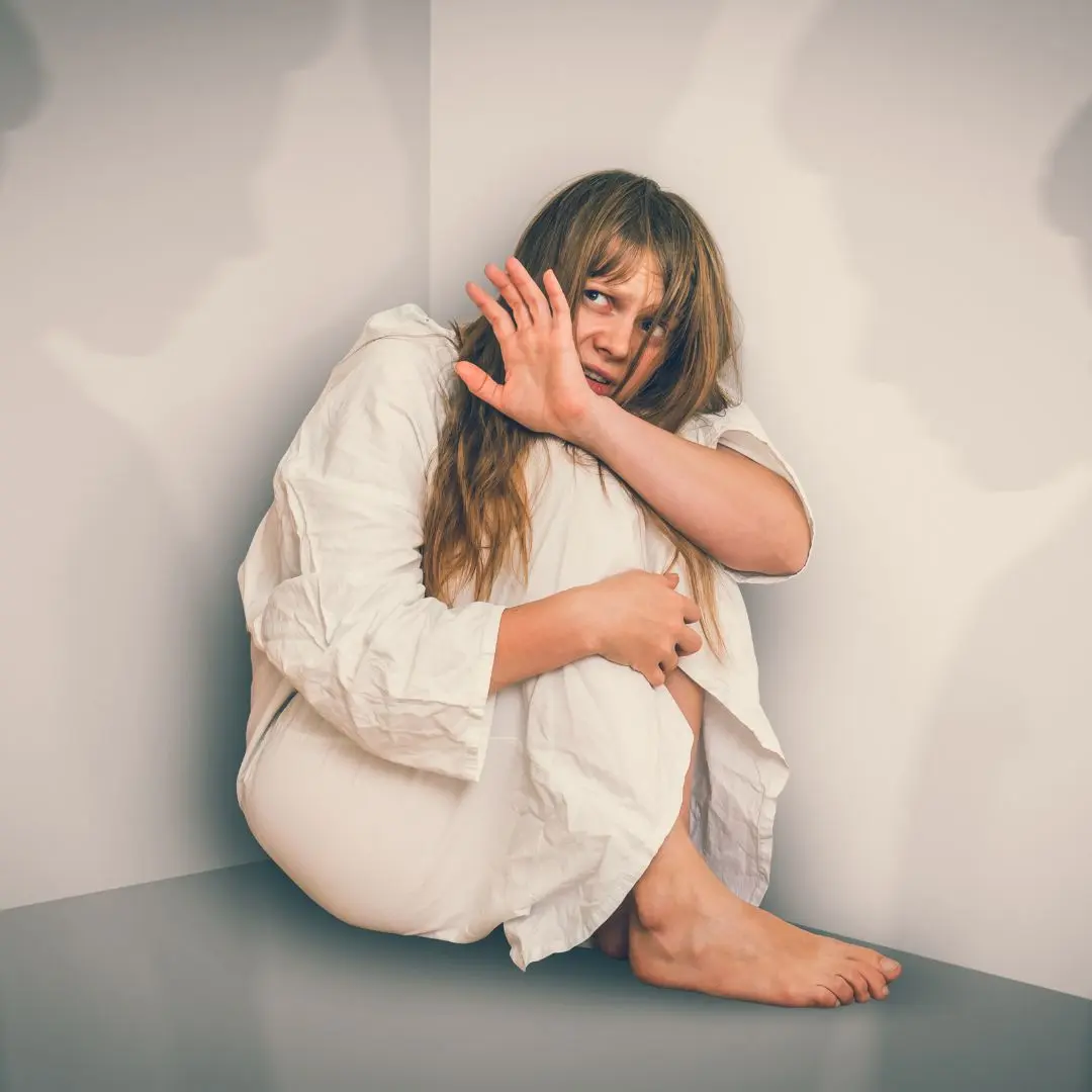 Young woman experiencing schizophrenia hallucinations, sitting in a corner with fear, illustrating the effects of severe mental health disorders.