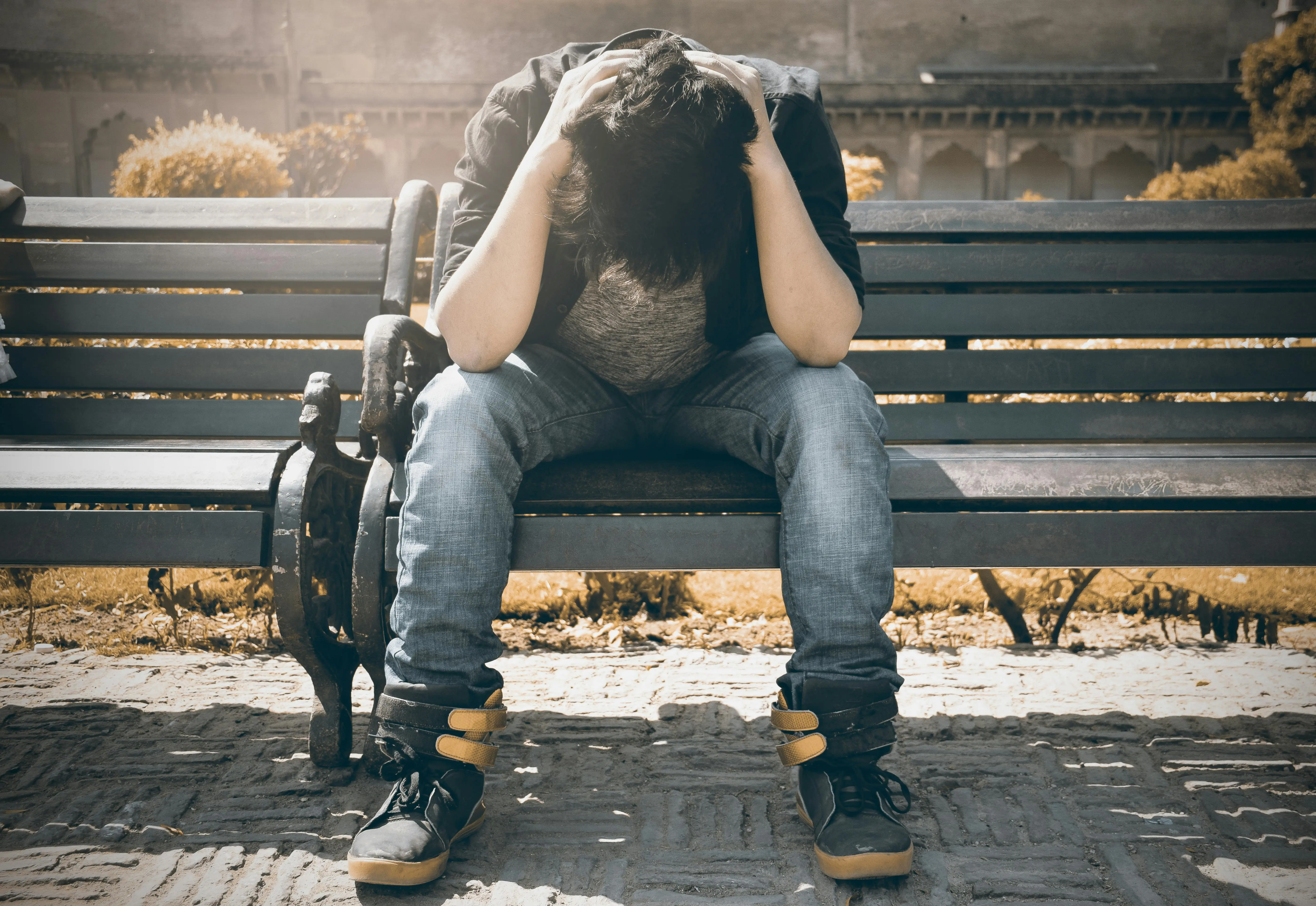 Stressed young man sitting on a park bench with head in hands, struggling with anxiety and mental health challenges.