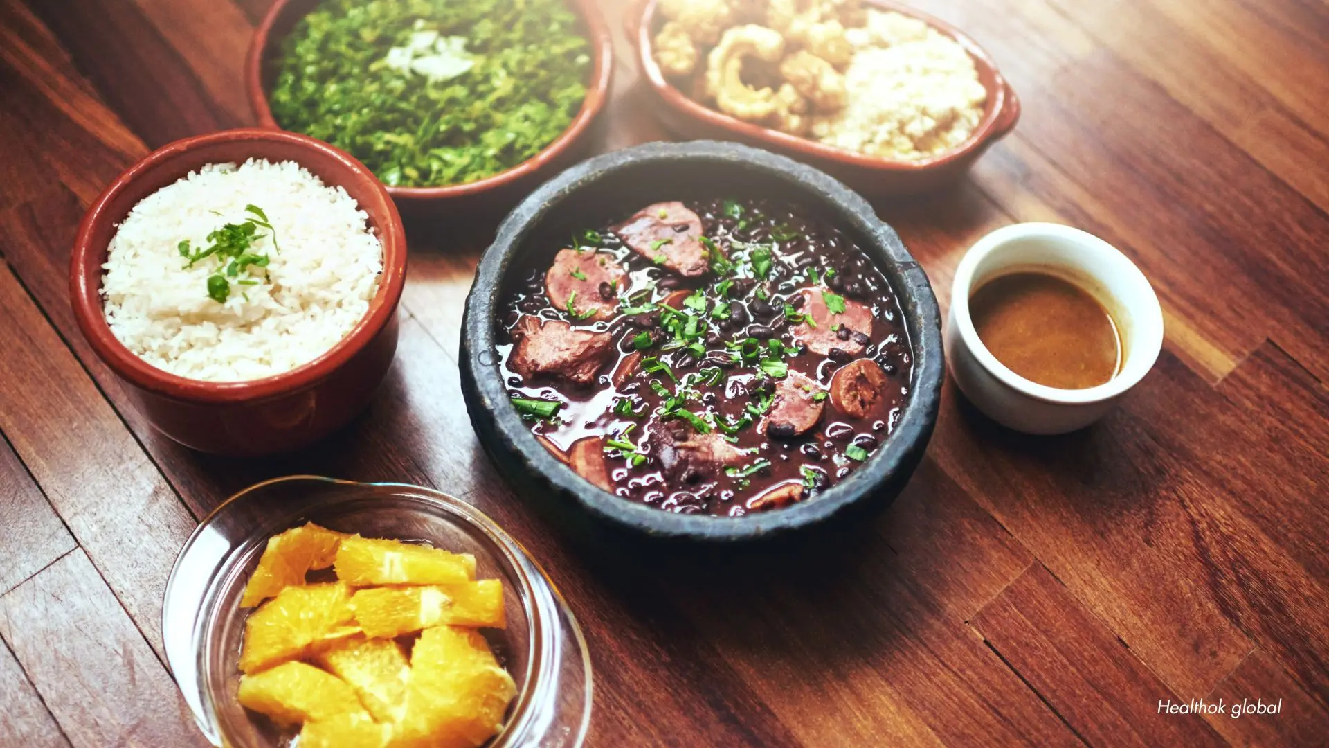 Traditional wholesome meal featuring a bowl of black bean stew with herbs, white rice, fresh orange slices, sautéed greens, and a cup of sauce, representing a balanced and nutrient-rich diet.